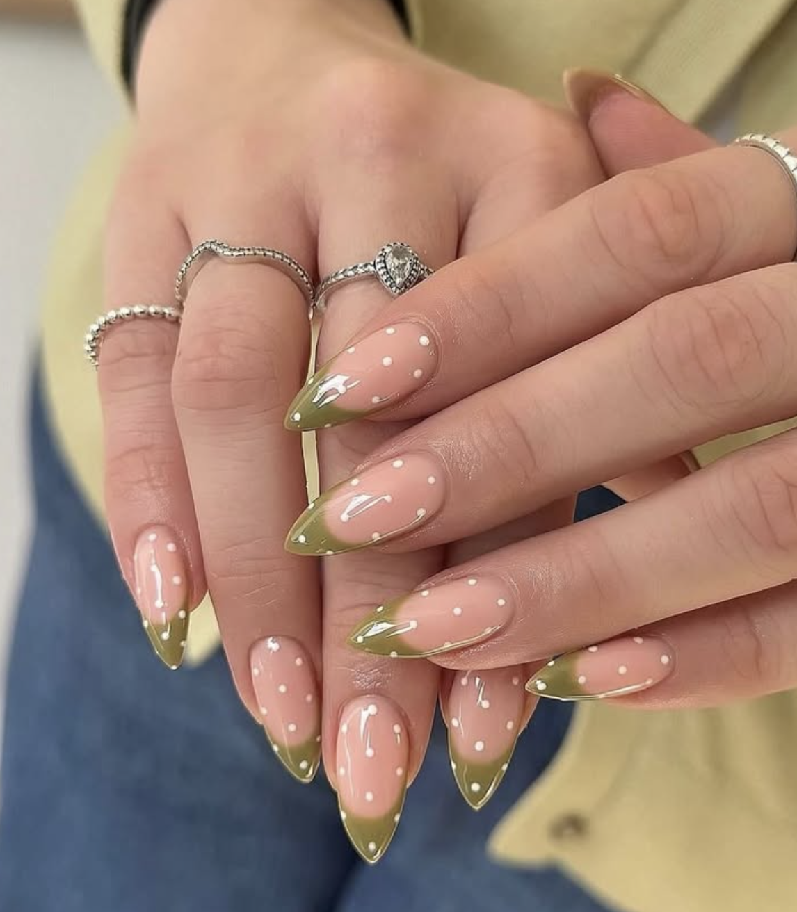 Close-up of almond-shaped nails featuring a muted olive green French tip design with delicate white polka dots and a high-shine finish.