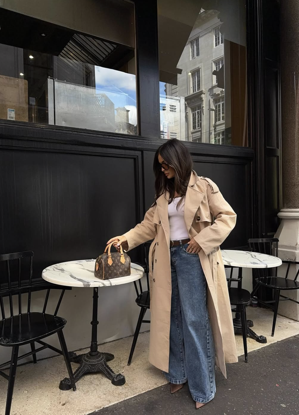 A woman standing outside a cafe wearing wide-leg blue jeans, a white tank top, a classic tan trench coat, sunglasses, and resting a Louis Vuitton Speedy bag on a marble bistro table.