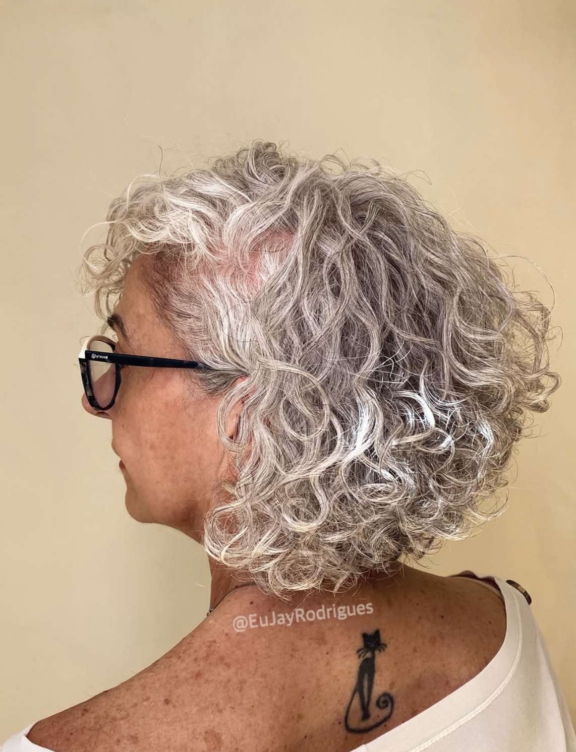 Side view of a woman over 65 wearing a rounded silver-gray curly bob with glasses.