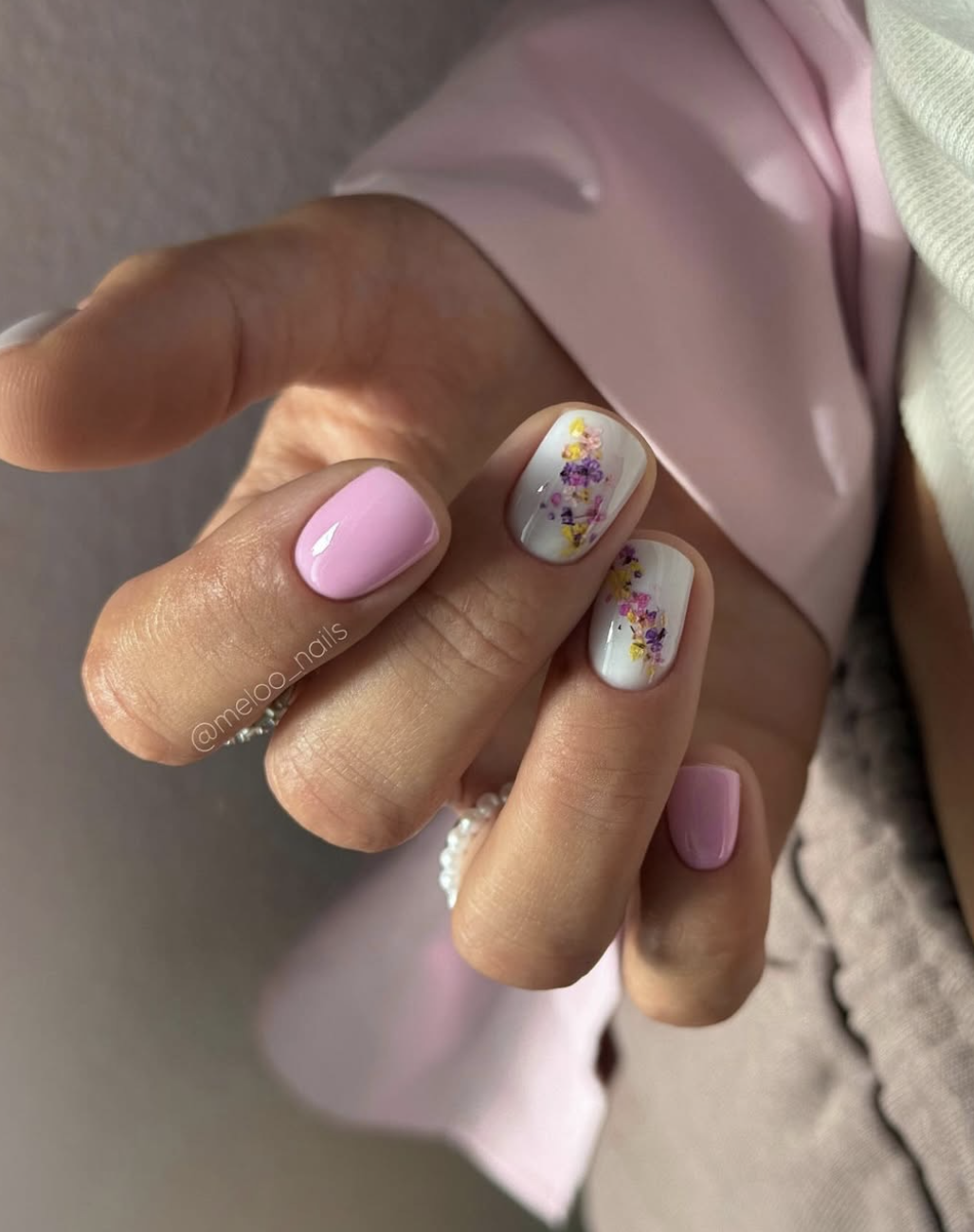 A close-up of short nails painted in light pink with delicate, hand-painted tiny floral nail art accents.