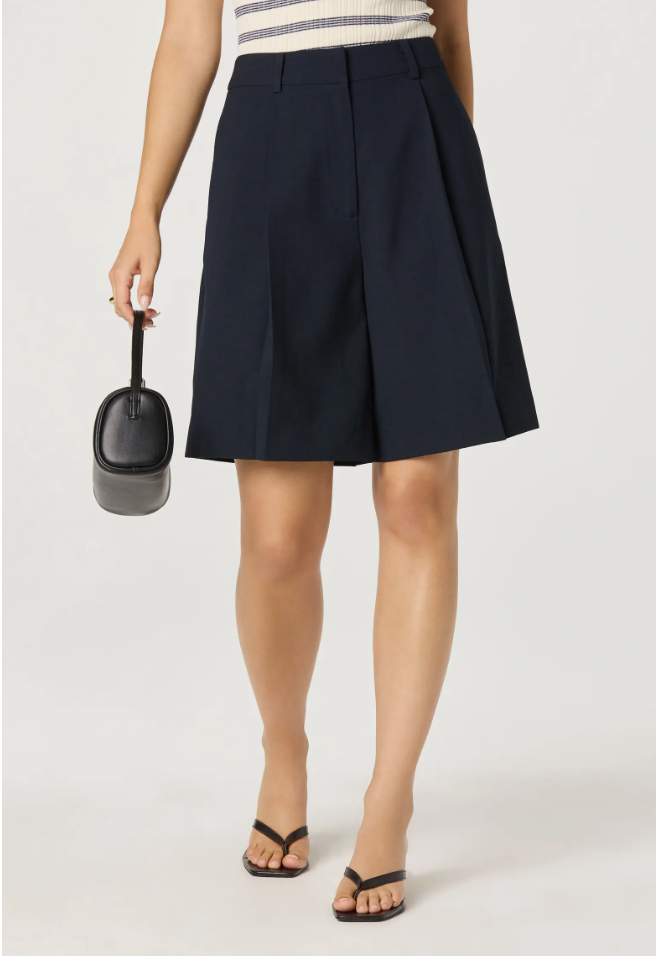 A close-up of a woman wearing navy blue tailored ASTR the Label Quillie pleated Bermuda shorts, styled with black thong sandals and a small black handbag.
