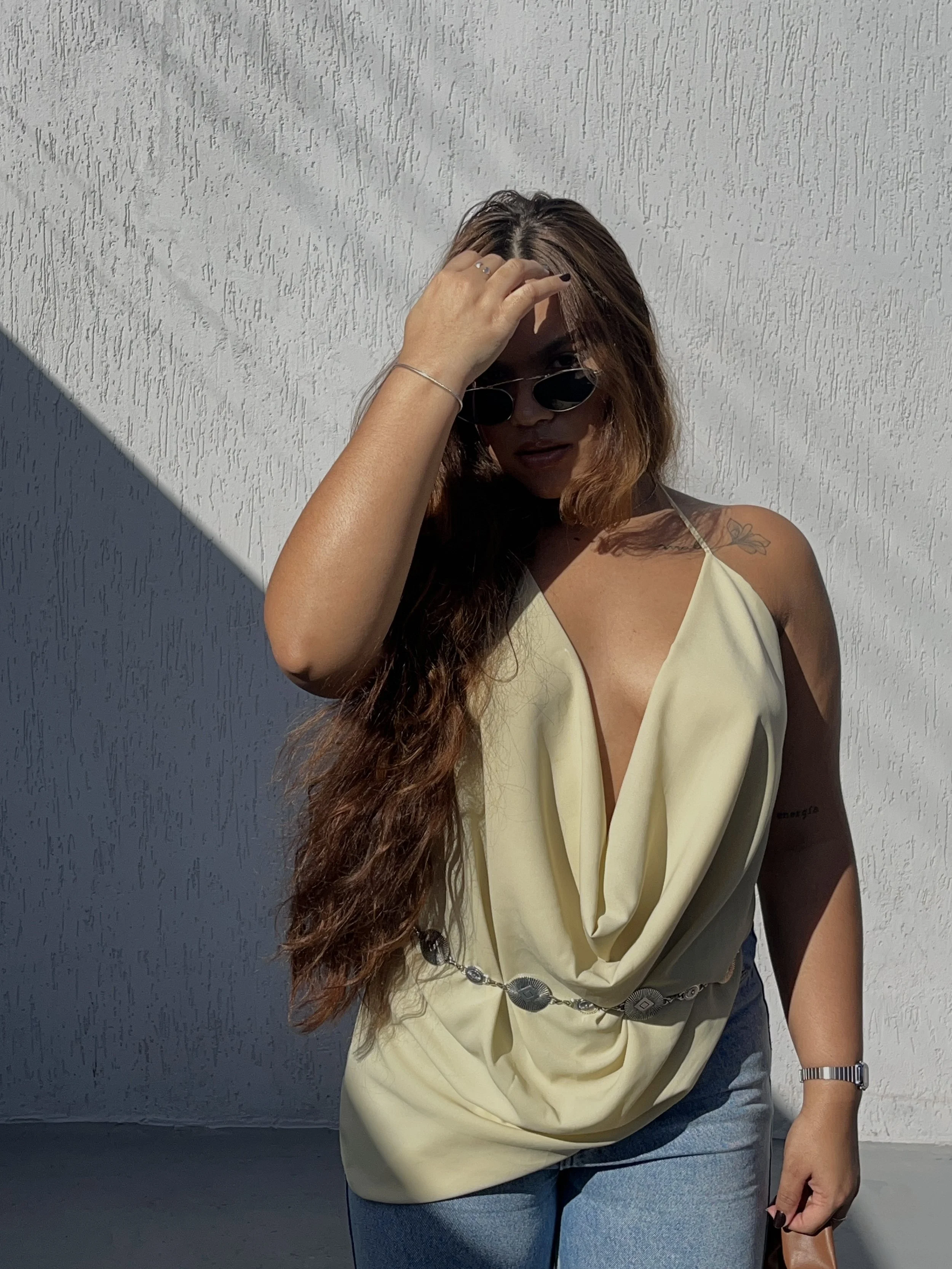 A woman with long, wavy brown hair wearing a draped cream cowl-neck halter top and a silver statement concho belt.