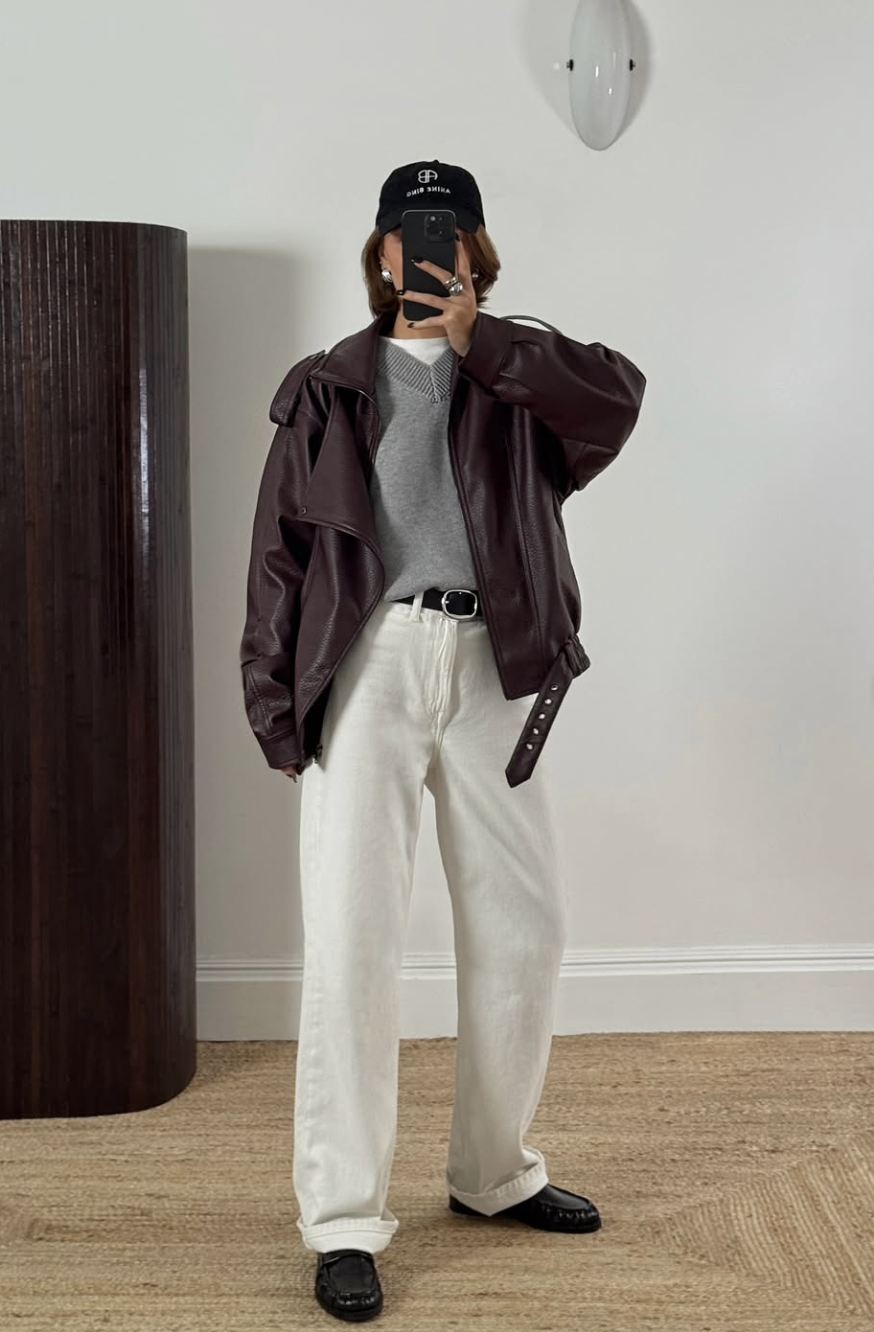 A woman wearing white straight-leg jeans with black pointed-toe loafers, a grey sweater, and an oversized burgundy leather bomber jacket.