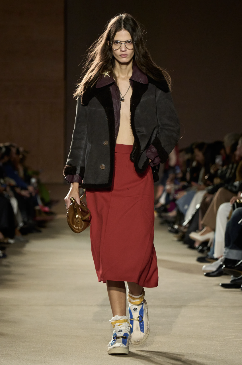 A model on the Coach Fall/Winter 2026 runway carrying a structured leather bag in a deep mahogany red, illustrating the season's muted clay color trend.