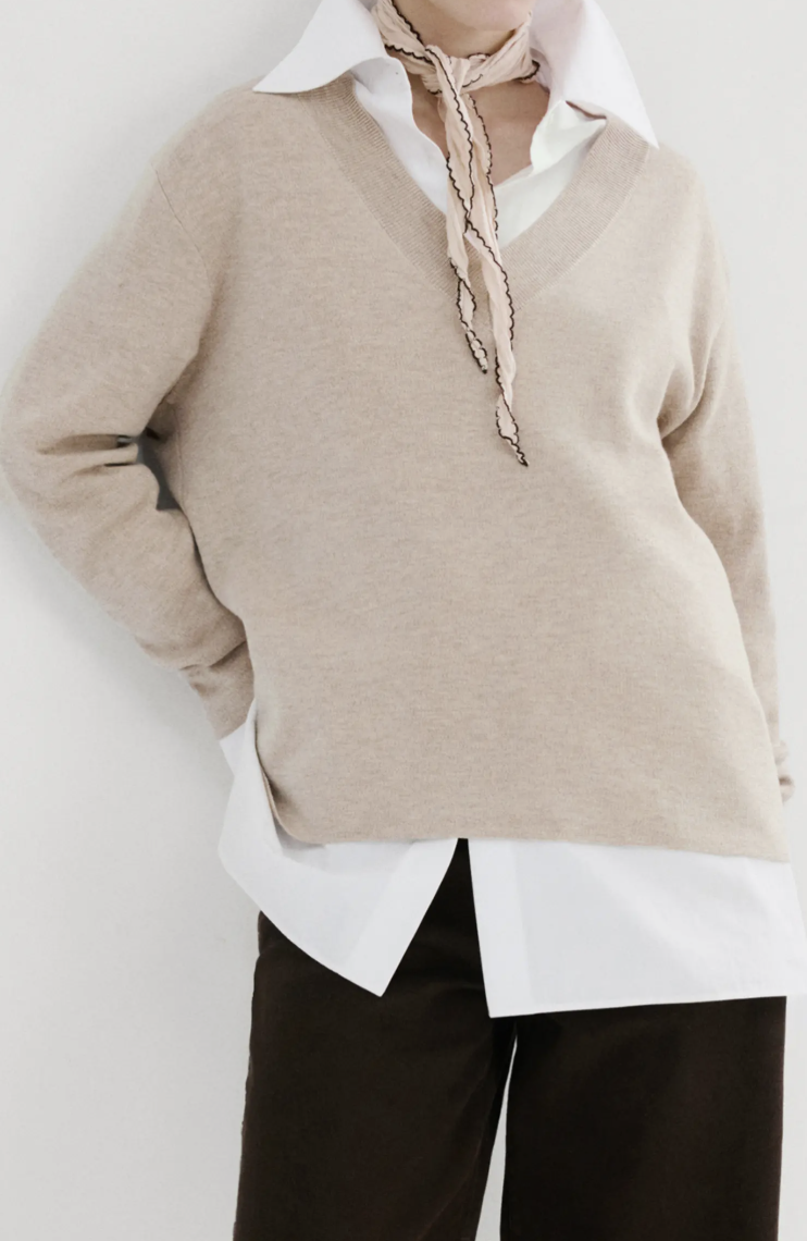 Woman wearing a beige V-neck sweater layered over a crisp white collared shirt, paired with dark green trousers for a preppy office outfit.