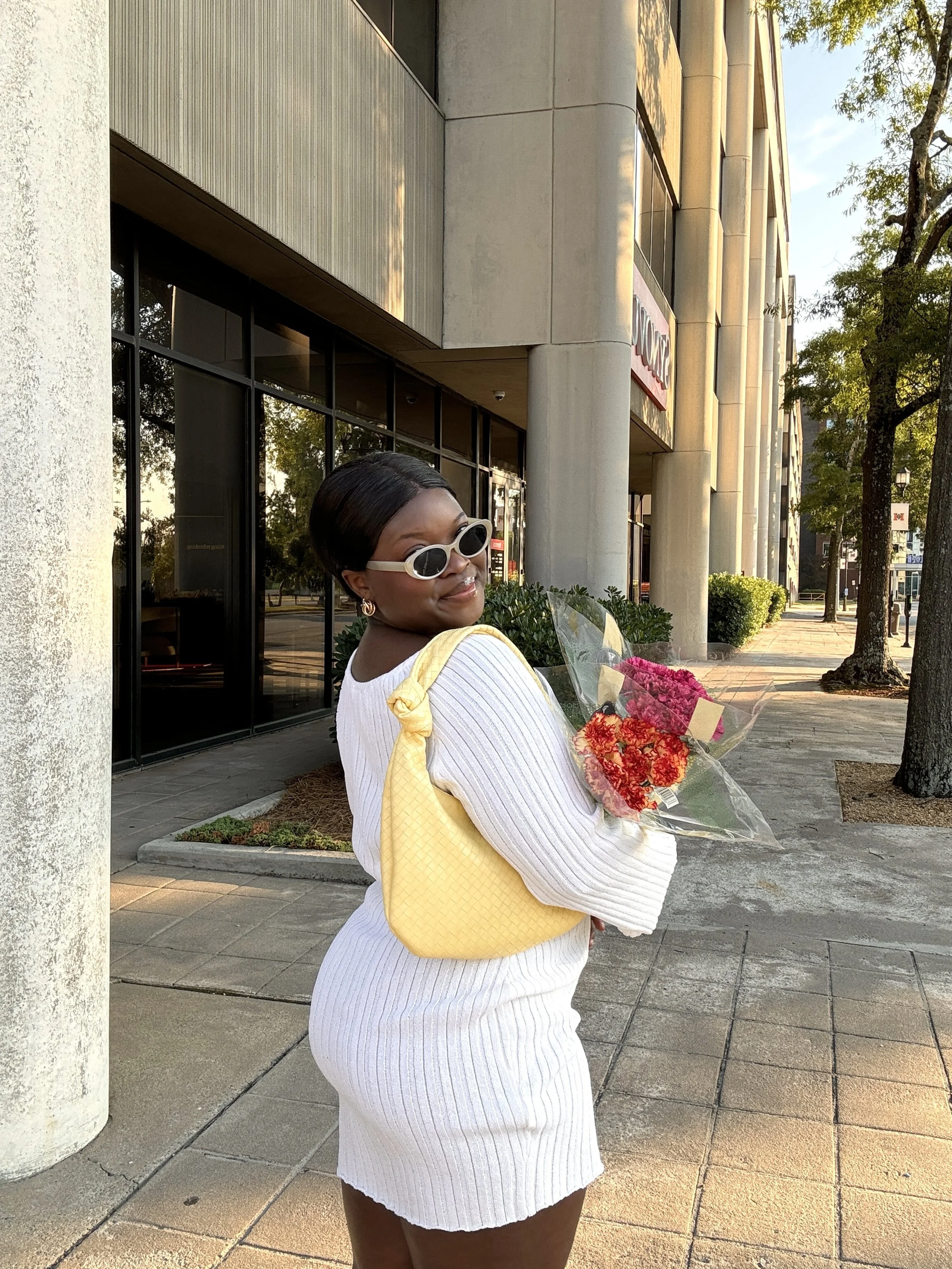 A woman wearing a white ribbed long-sleeved mini dress, retro white sunglasses, and a woven yellow shoulder bag, holding a bouquet of flowers.