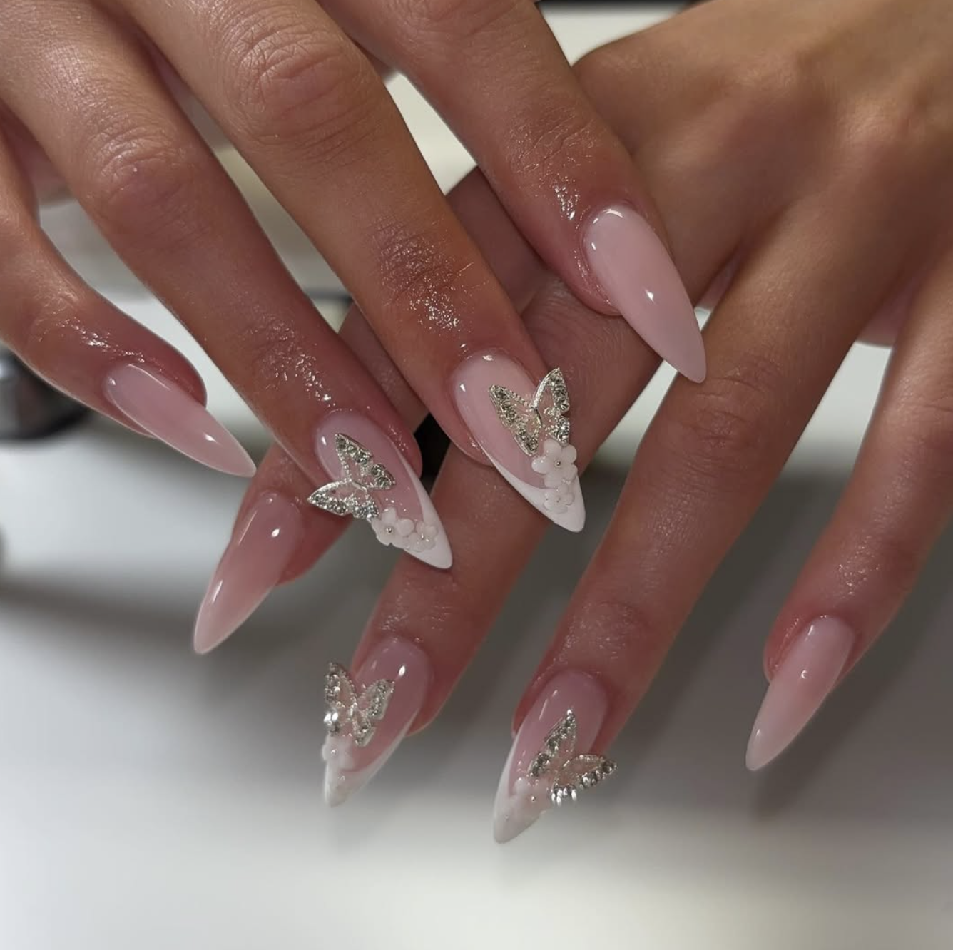 Long almond-shaped clear gel nails featuring 3D silver butterfly charms, tiny white flowers, and sparkling glitter accents.