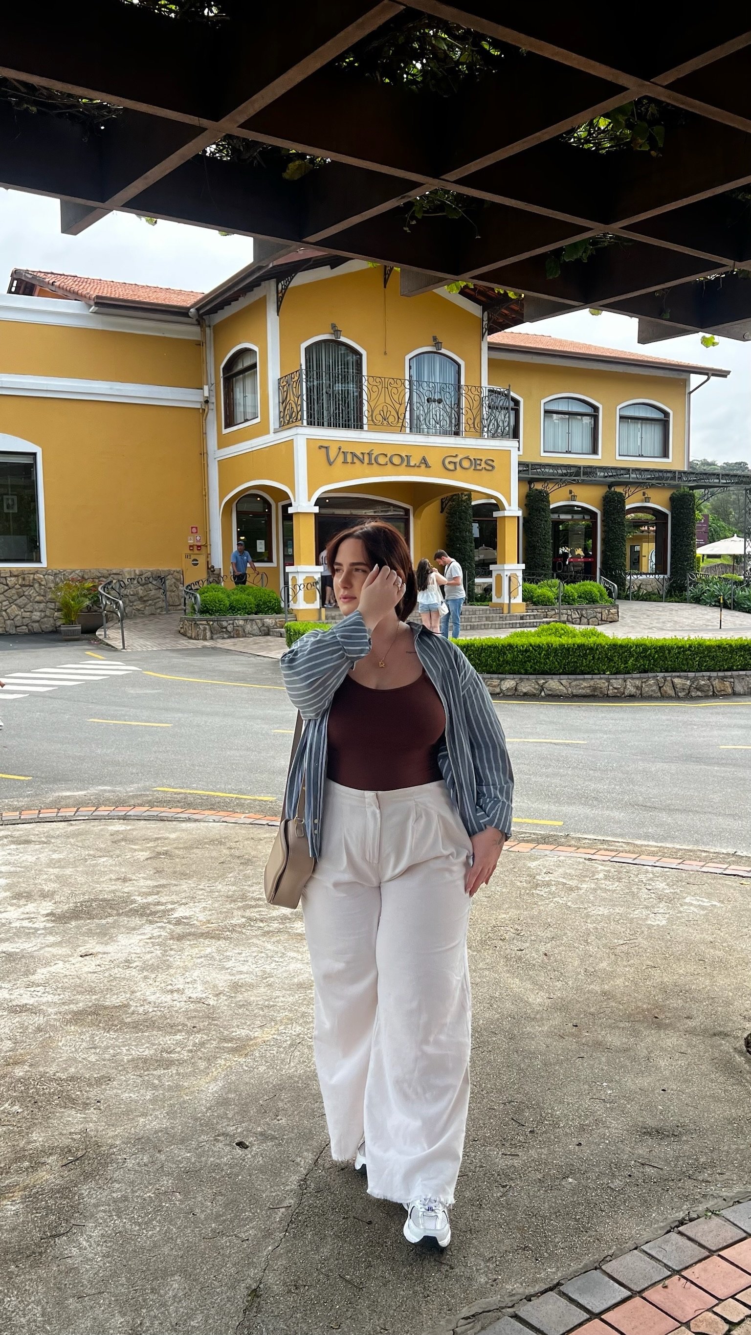 A woman wearing a blue and white striped button-down shirt over a white top with wide-leg white linen pants, standing in front of a yellow Mediterranean-style building.