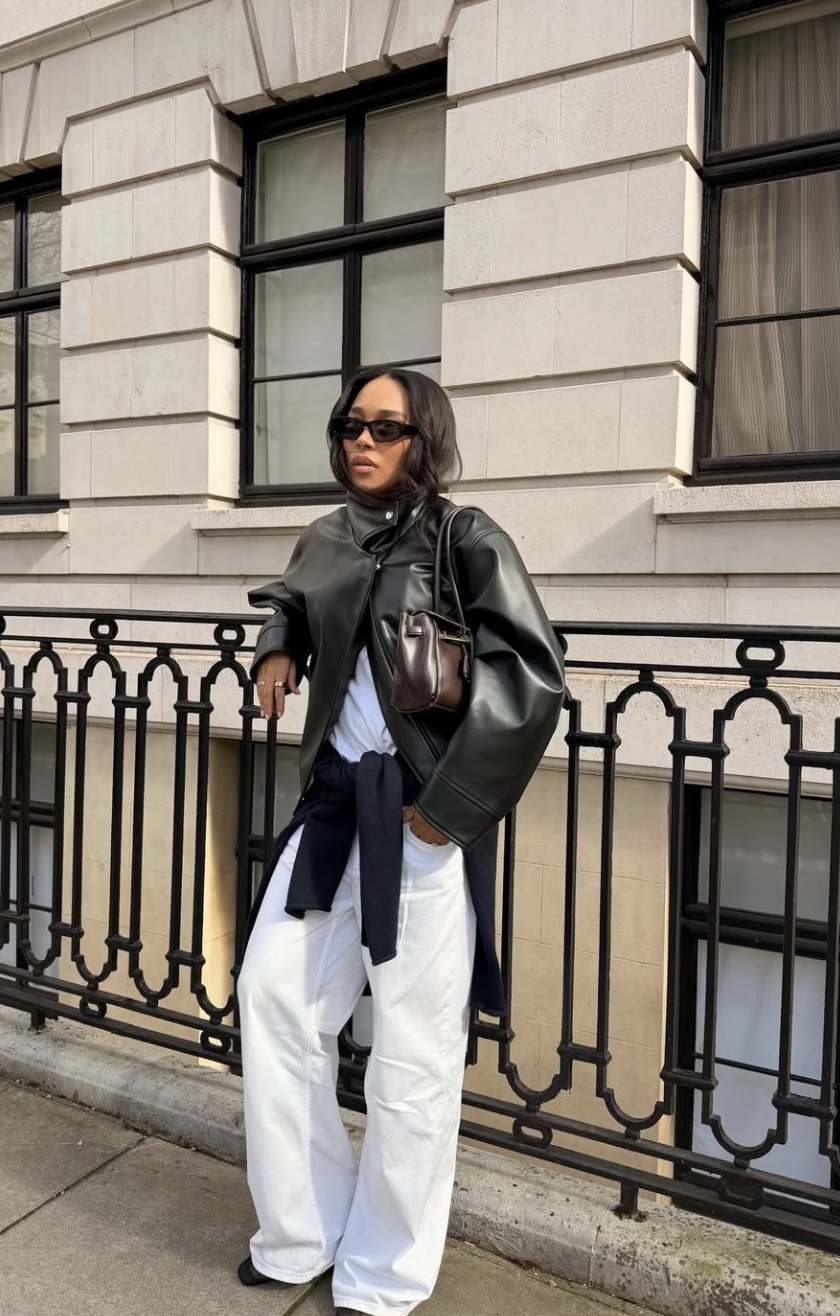 A woman wearing white straight-leg jeans with a black oversized leather jacket, black pointed-toe mules, and a dark brown shoulder bag.