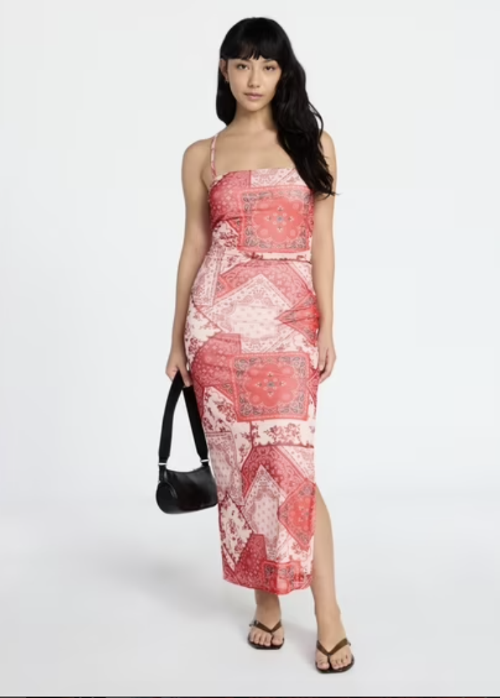 A full-length product shot of a high-waisted bohemian maxi skirt featuring a patchwork design of pink and red paisley patterns, a side thigh slit, styled with a black leather shoulder bag and brown thong sandals.