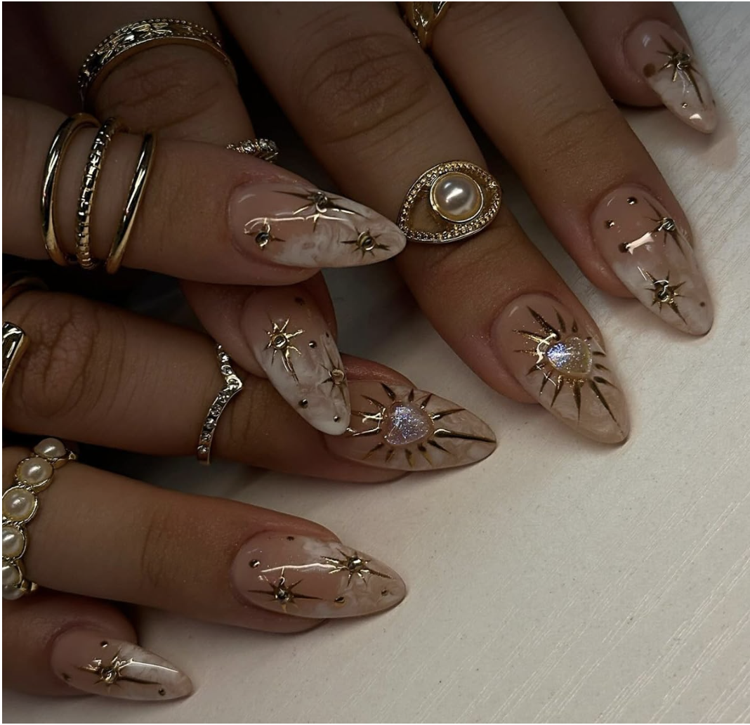 Almond-shaped press-on nails with a nude base featuring 3D gold boho stars, celestial sunbursts, and small heart charms.