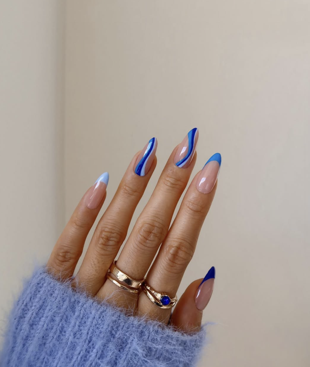 A close-up of long almond-shaped nails with an ocean-blue French manicure and abstract wavy line nail art, paired with a fuzzy blue sweater.