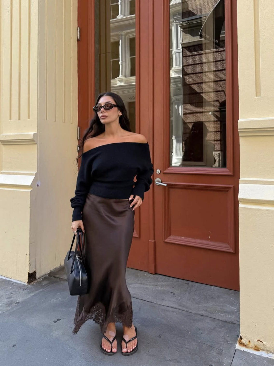 Woman wearing a brown satin lace-trim midi skirt with a navy off-the-shoulder sweater and black thong sandals
