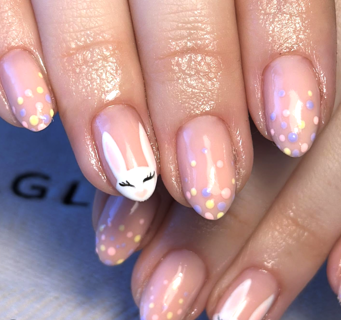 A close-up of almond-shaped nails with a sheer nude base, featuring a hand-painted white bunny with black eyelashes and various pastel polka dots in lavender, mint, and yellow.