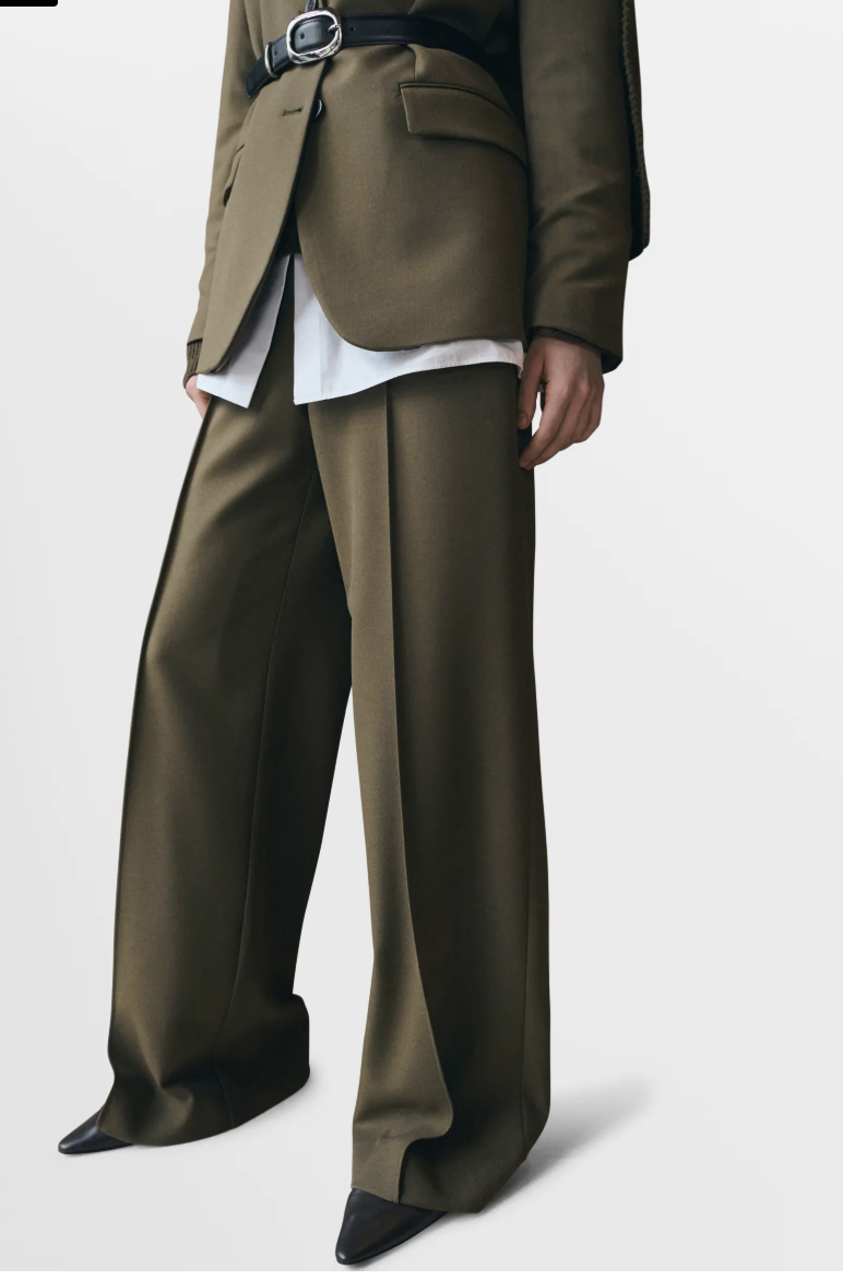 Close up of olive green pleated straight-leg trousers paired with a black top and black pointed-toe boots.