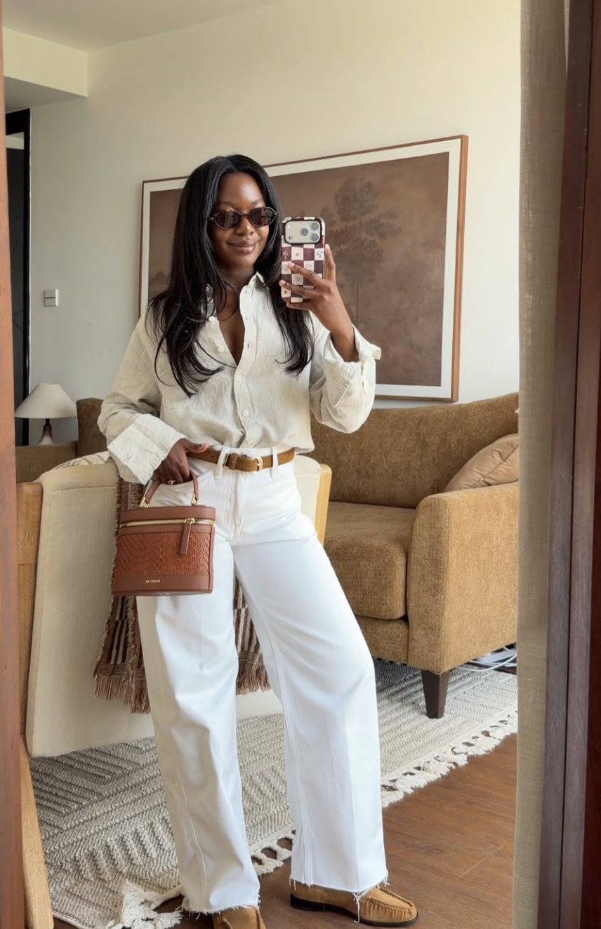 A woman taking a mirror selfie wearing white wide-leg jeans, a tucked-in linen button-down shirt, a brown leather belt, and a matching woven handbag.