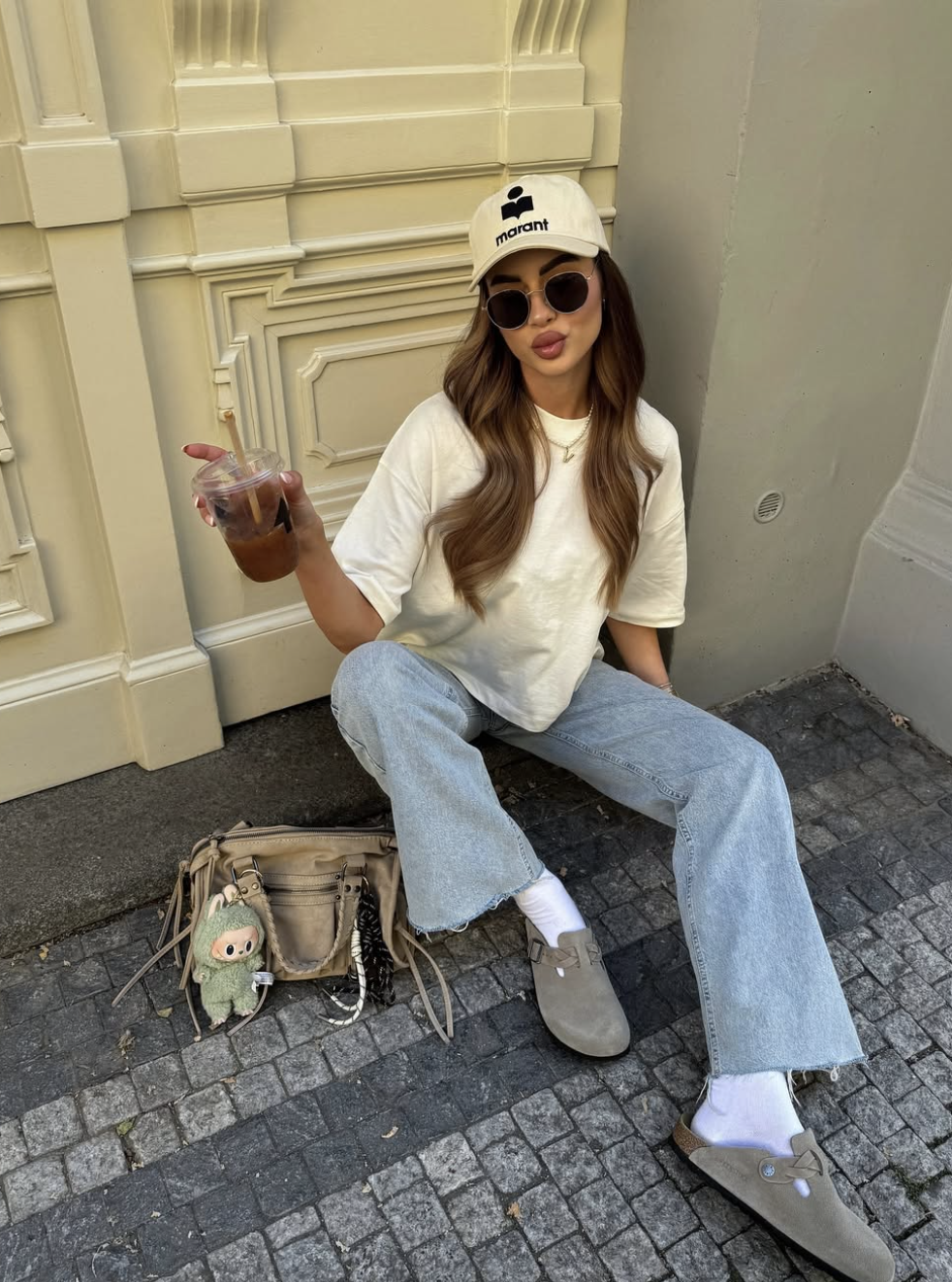 A woman sitting on a cobblestone street wearing a cream oversized t-shirt, light-wash frayed jeans, white socks, and grey suede clogs, holding an iced coffee next to a tan utility shoulder bag.