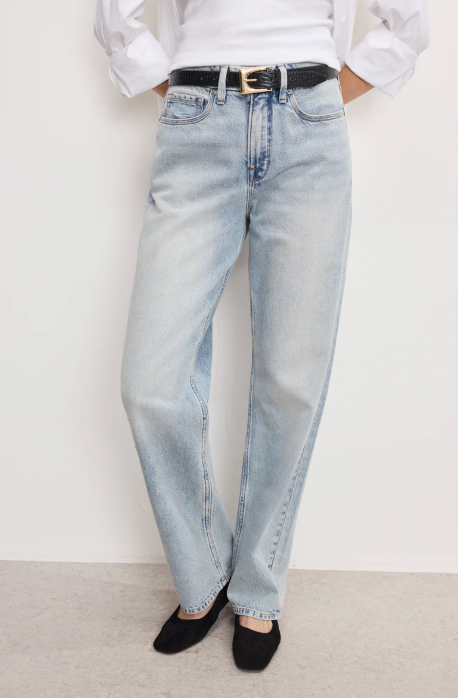A close-up style shot of a woman wearing light wash straight-leg denim jeans paired with a tucked-in white button-down shirt and a black textured leather belt with a gold buckle.