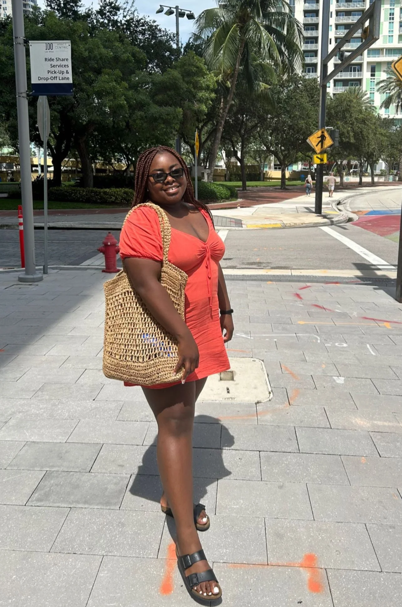 A woman standing outdoors wearing a vibrant orange puff-sleeve mini dress, a woven straw tote bag, and black mesh Mary Jane ballet flats, a key footwear trend for Spring 2026.