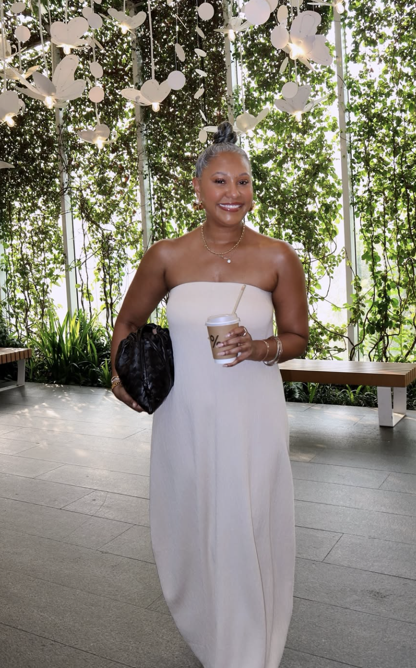 A woman wearing an elevated monochromatic cream pleated outfit featuring a matching neck scarf, a dark chocolate brown suede tote bag, and tortoiseshell sunglasses.