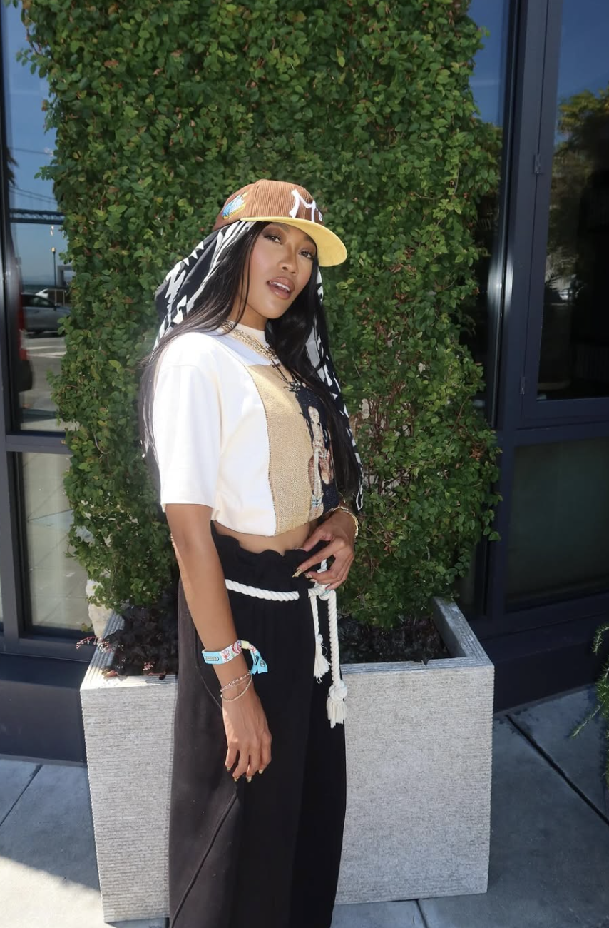 A woman posing in front of a green ivy wall wearing a brown and yellow baseball cap over a black and white patterned headscarf, a cream graphic t-shirt, and black high-waisted pants with a thick white rope belt.