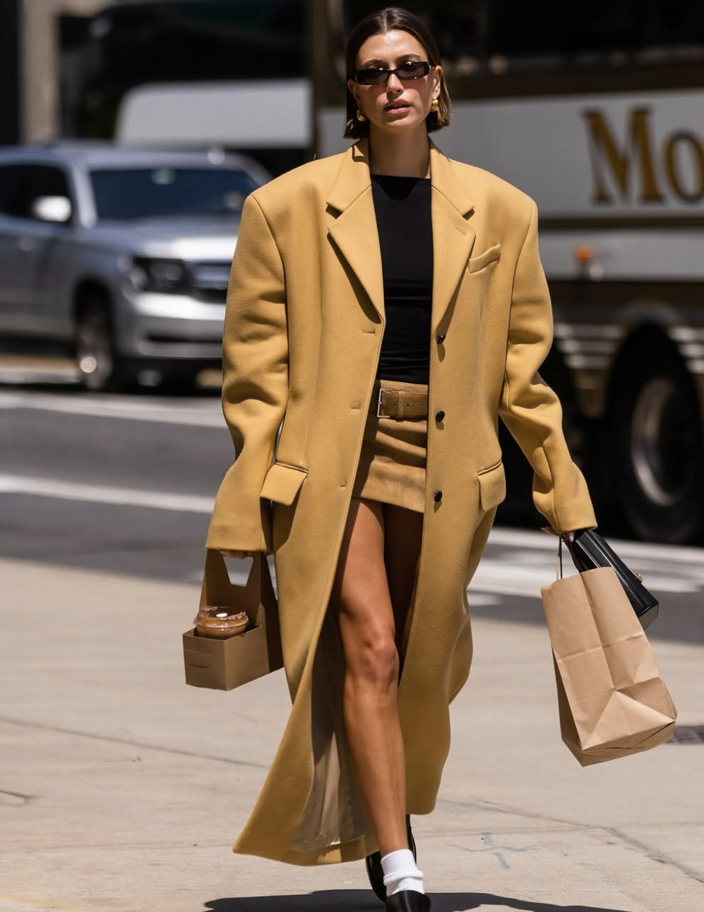Hailey Bieber walking on a city street wearing a long tan trench coat, a black mini skirt, white ruffled lace ankle socks, and black leather loafers.