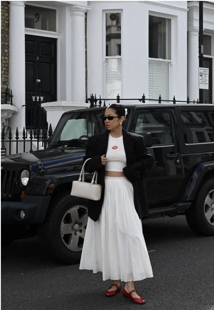A woman wearing an oversized blazer, a cropped graphic tee, and a white pleated maxi skirt paired with black Mary Jane flats for women.