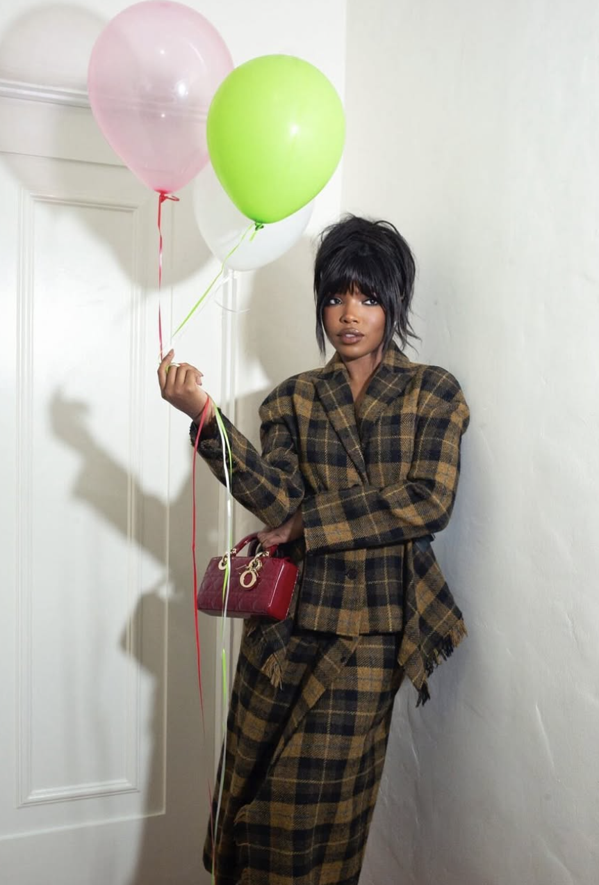 Ryan Destiny wearing a plaid blazer and holding balloons, featuring a high messy bun with piecey fringe bangs that soften the forehead and balance a heart face shape.