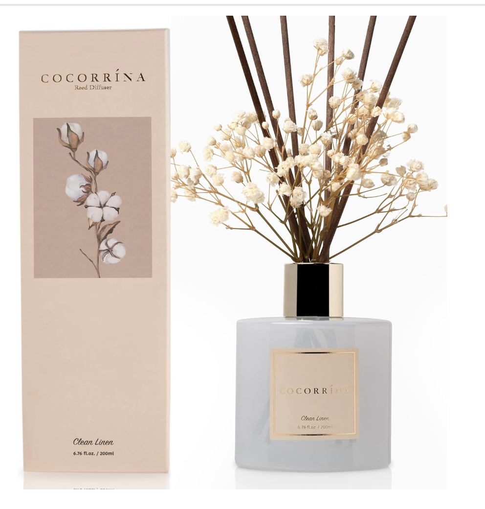 A Cocorrina Clean Linen reed diffuser set featuring a white glass bottle with a gold accent ring, reed diffusers with floral accents, and matching cream packaging.