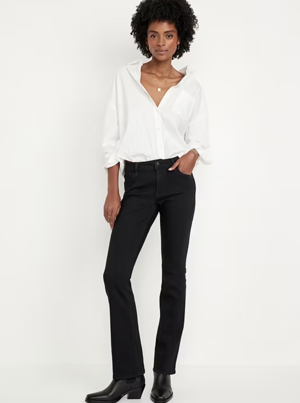 A person with an hourglass figure wearing black Old Navy Mid-Rise Wow Boot-Cut jeans and black boots, showing the subtle flare at the hem and the contouring fit through the hips.