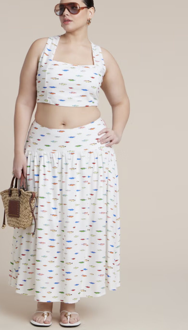 Plus-size woman wearing a white matching co-ord set with a colorful fish print, featuring a square-neck crop top and a tiered maxi skirt while holding a small woven tote bag.