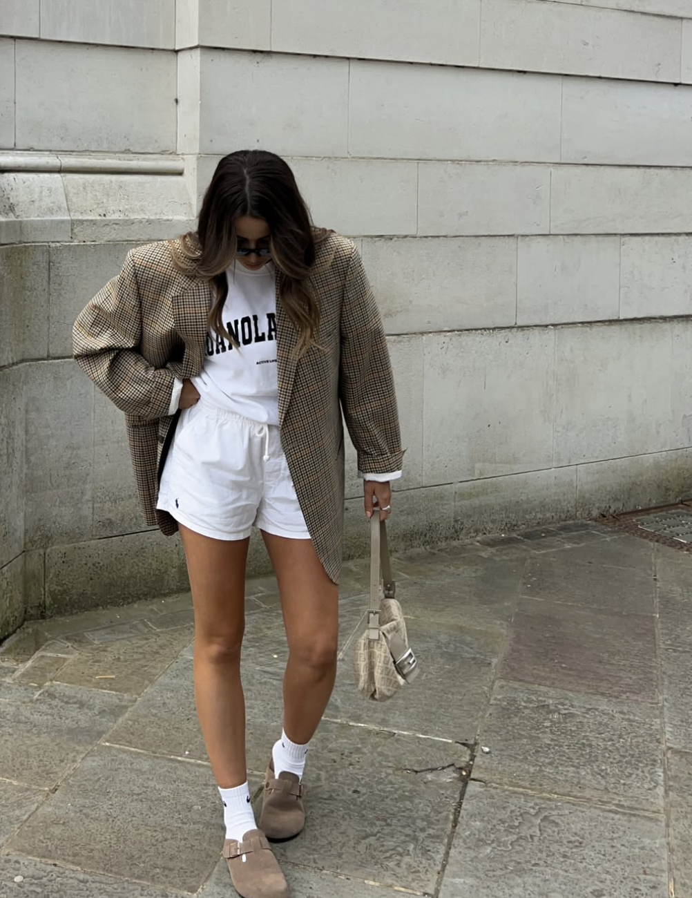A woman wearing an oversized brown houndstooth blazer over a white graphic t-shirt and matching white track shorts, accessorized with a plush shoulder bag for a chic summer street style look.