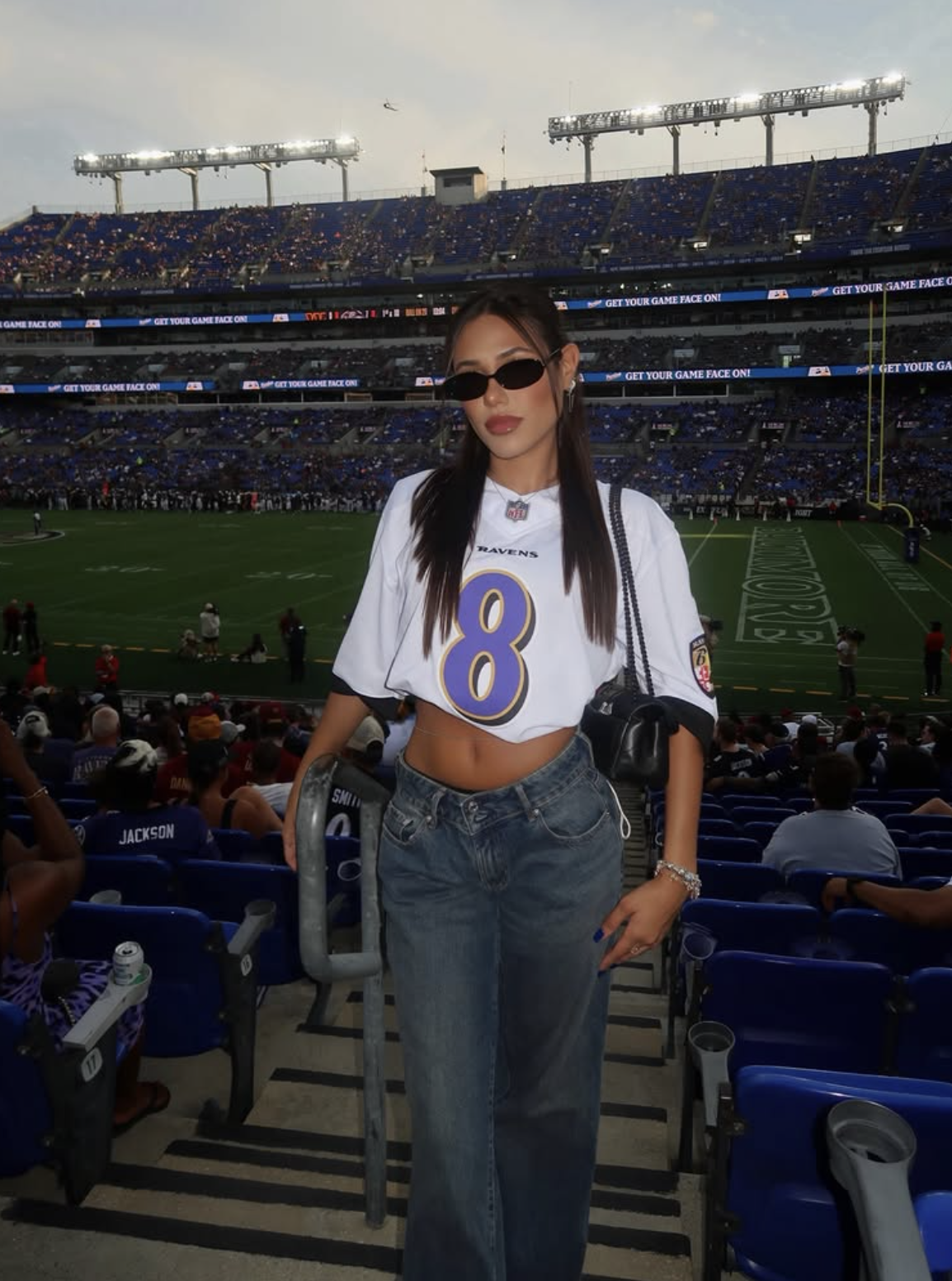 Woman wearing an oversized white Baltimore Ravens jersey with relaxed dark-wash jeans, sunglasses and a small black bag.