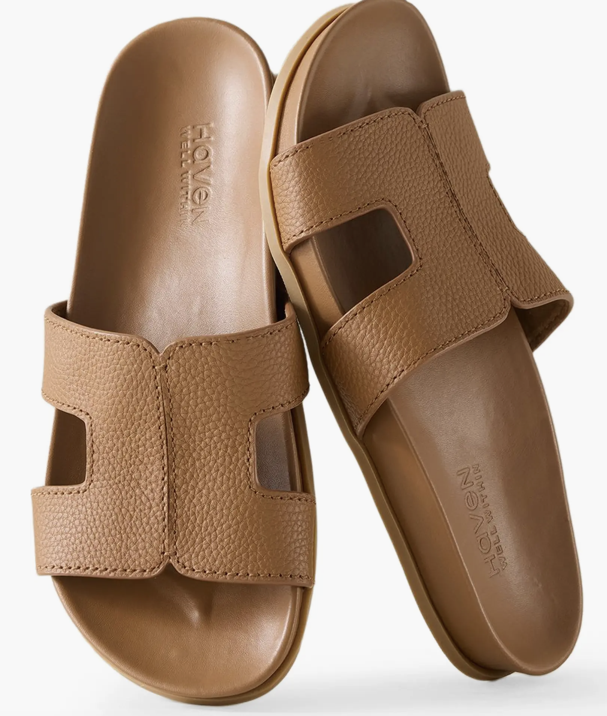 A close-up of a pair of tan pebble-grain leather slide mules featuring a wide cross-strap design and a comfortable molded footbed.