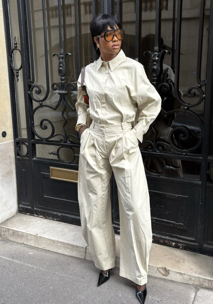 A woman with a pixie cut haircut wearing large orange sunglasses and a monochromatic beige button-down shirt and matching wide-leg pleated trousers.