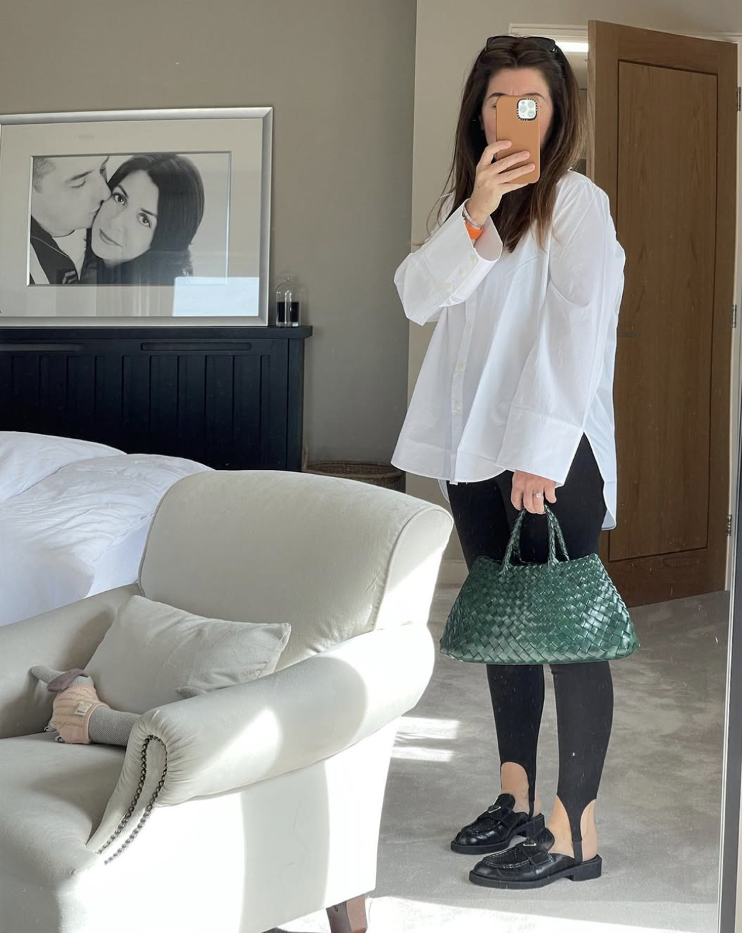 A woman taking a mirror selfie wearing an oversized white button-down shirt, black leggings, and chunky black loafers, carrying a large green woven tote bag.