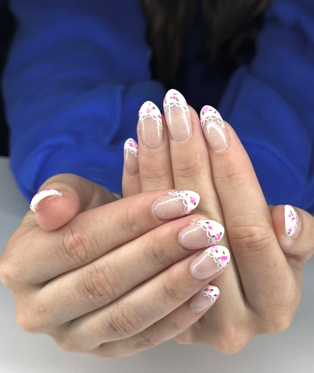 lose-up of a hand with a "vintage lace" manicure on almond-shaped nails, featuring delicate white lace patterns on the tips and tiny hand-painted pink rosebuds with green leaves.