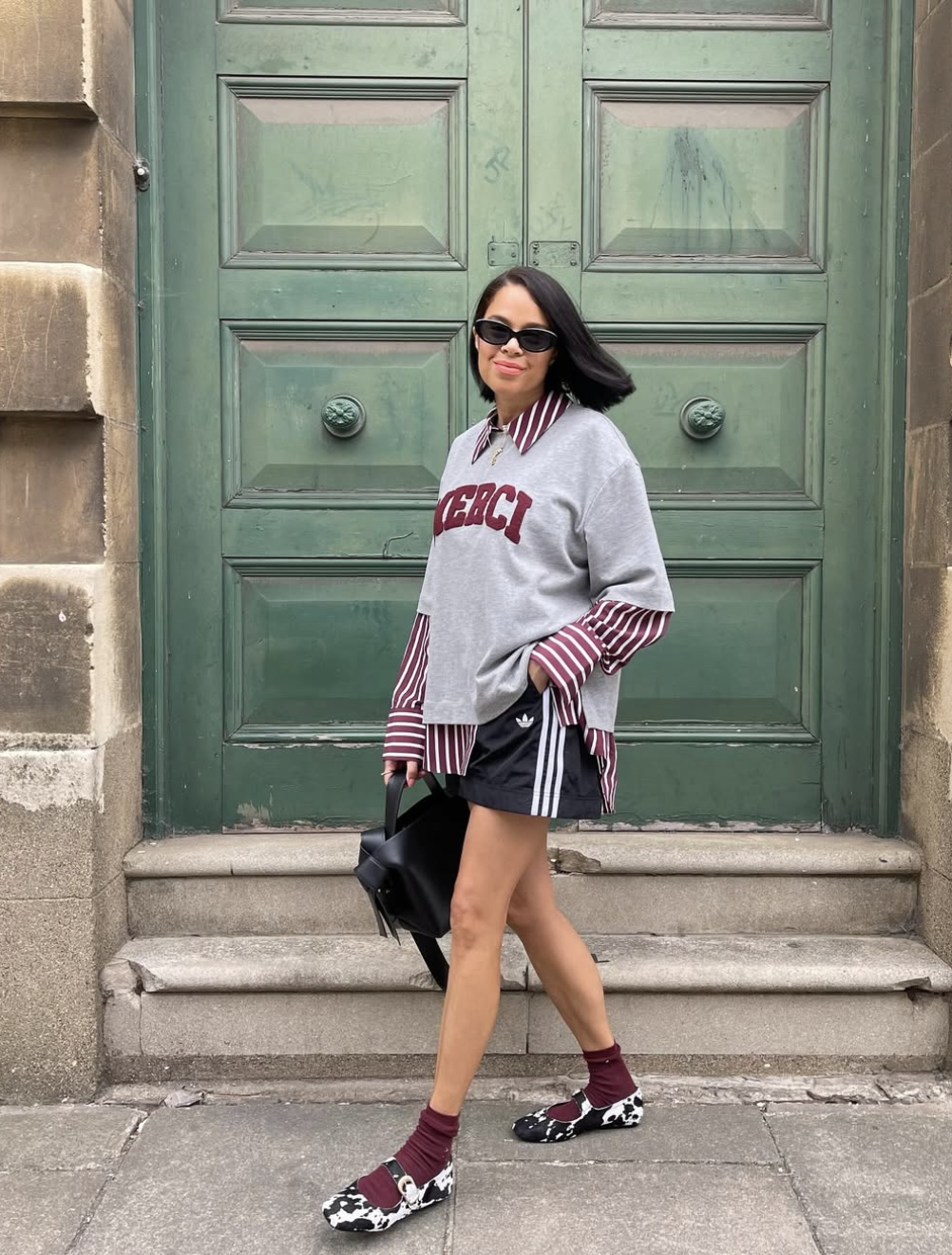 A woman walking in front of a green door wearing a grey crewneck sweatshirt layered over a burgundy striped button-down shirt, black Adidas track shorts, maroon socks, and patterned loafers.