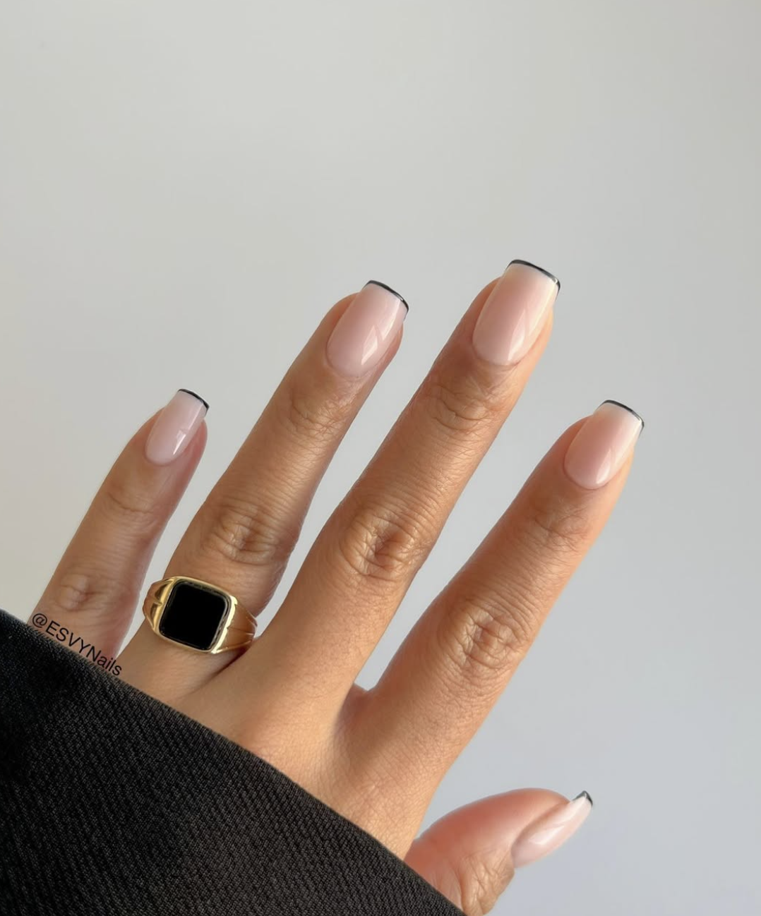 A hand with short nails featuring a thin, micro French tip manicure with a sheer nude base and gold ring accent.