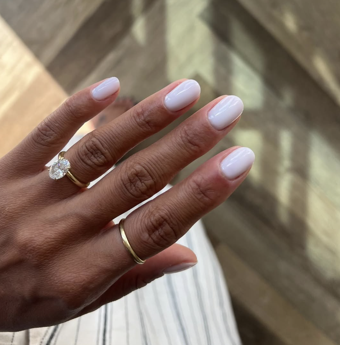 A close-up of short, round-shaped nails with a glossy, semi-translucent coconut milk white manicure on a person with a deep skin tone, wearing a gold wedding band and a large oval diamond engagement ring.