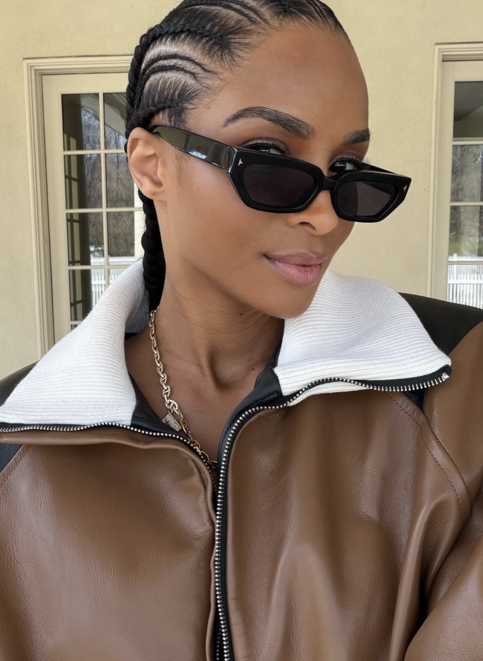 Ciara wearing a brown leather jacket and black sunglasses, showcasing sleek straight-back cornrows with long braided ends that create an elongating effect for a round face shape.