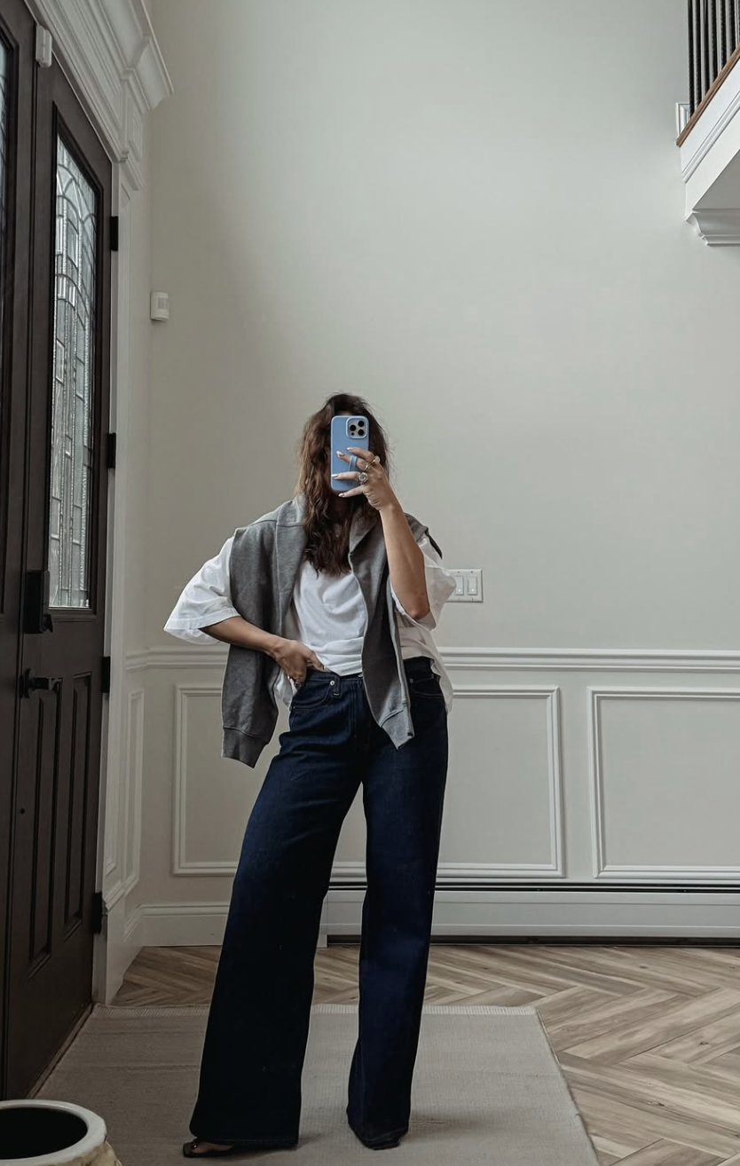 A woman wearing a white t-shirt and dark wash jeans with a grey crewneck sweatshirt draped over her shoulders, styled as a chic and casual everyday outfit.