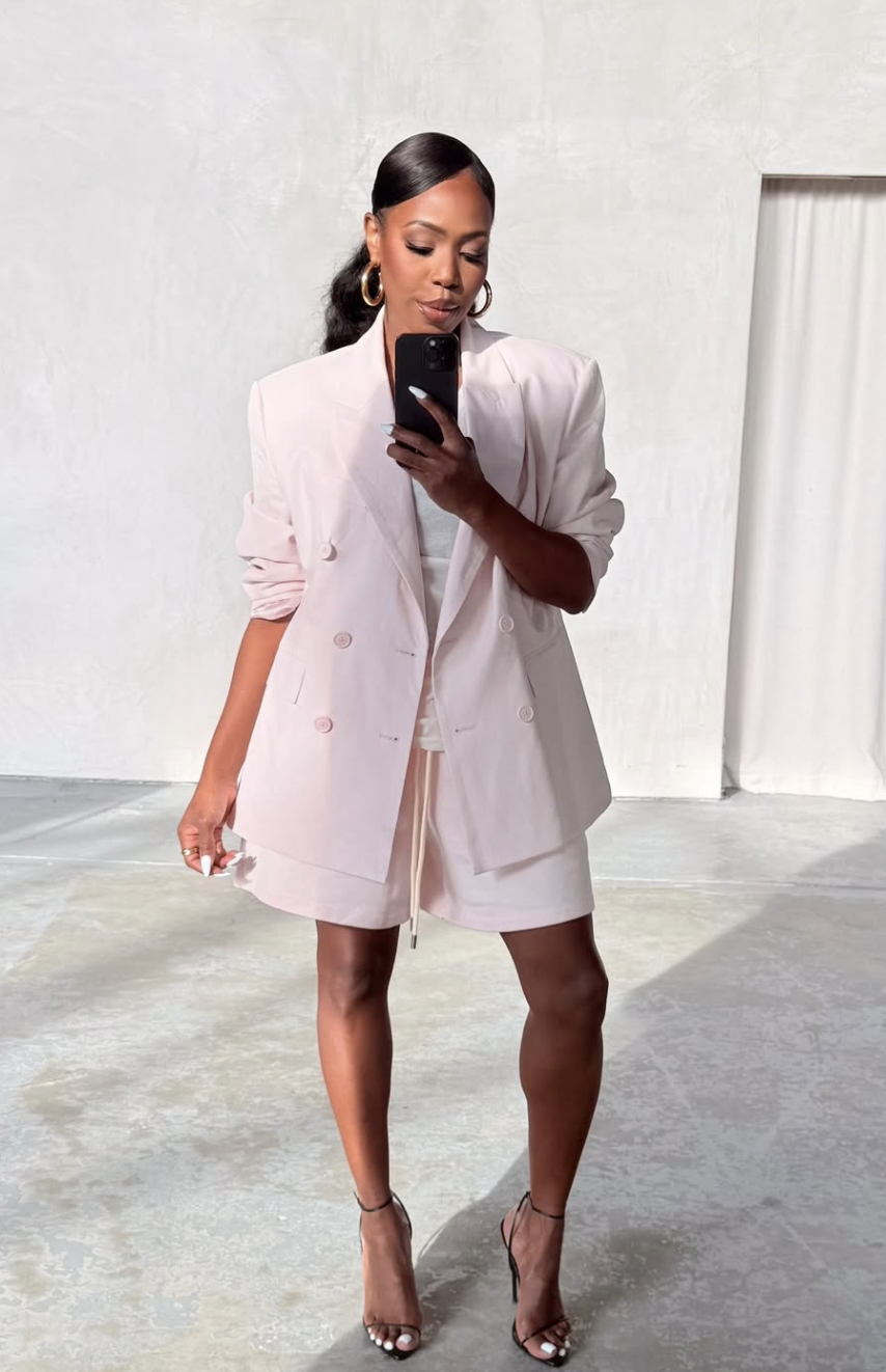 A woman wearing a monochromatic soft pink outfit featuring an oversized blazer, a matching top, and shorts, styled with gold hoop earrings.
