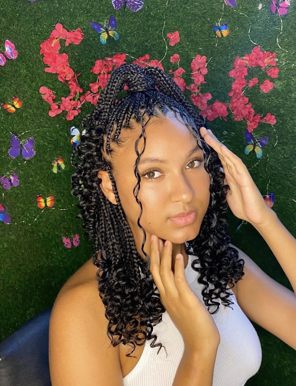 A woman with short, shoulder-length knotless braids and a braided fringe, featuring curly face-framing tendrils and a half-up ponytail against a floral butterfly background.