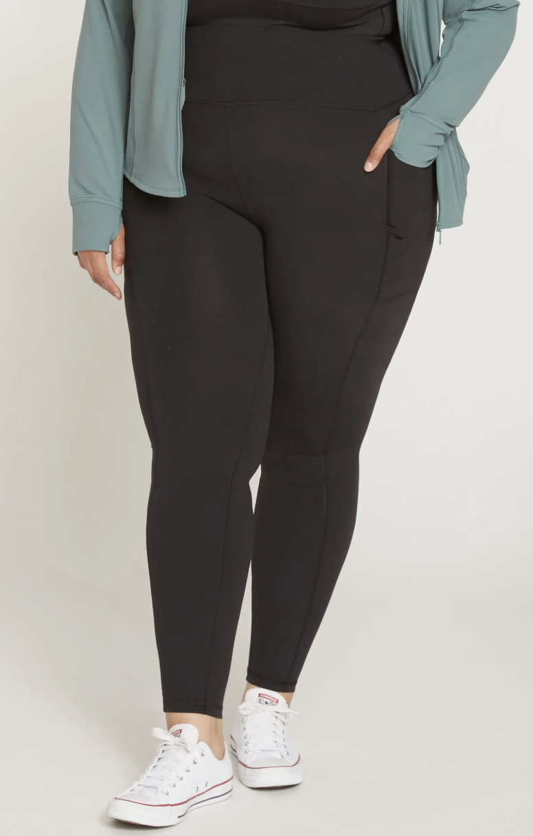 A close-up shot of a plus-size model wearing high-waisted black performance leggings styled with white Converse sneakers and a light blue athletic jacket, demonstrating a practical and polished everyday outfit for 2026.