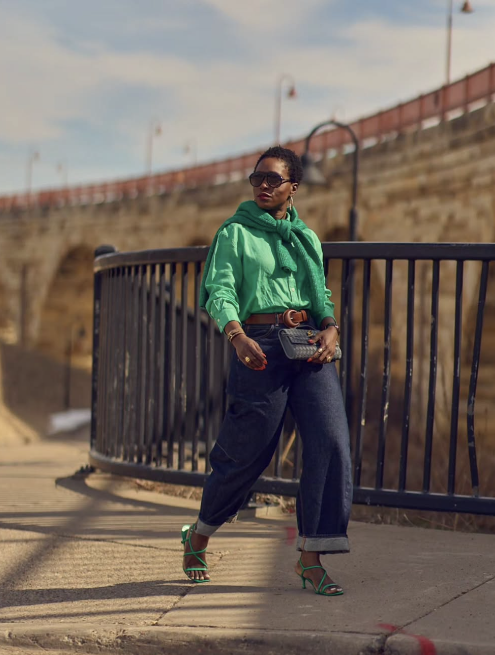 A woman wearing a vibrant green satin button-down shirt with a matching green sweater draped over her shoulders, paired with dark wash wide-leg cuffed jeans and green strappy heels.