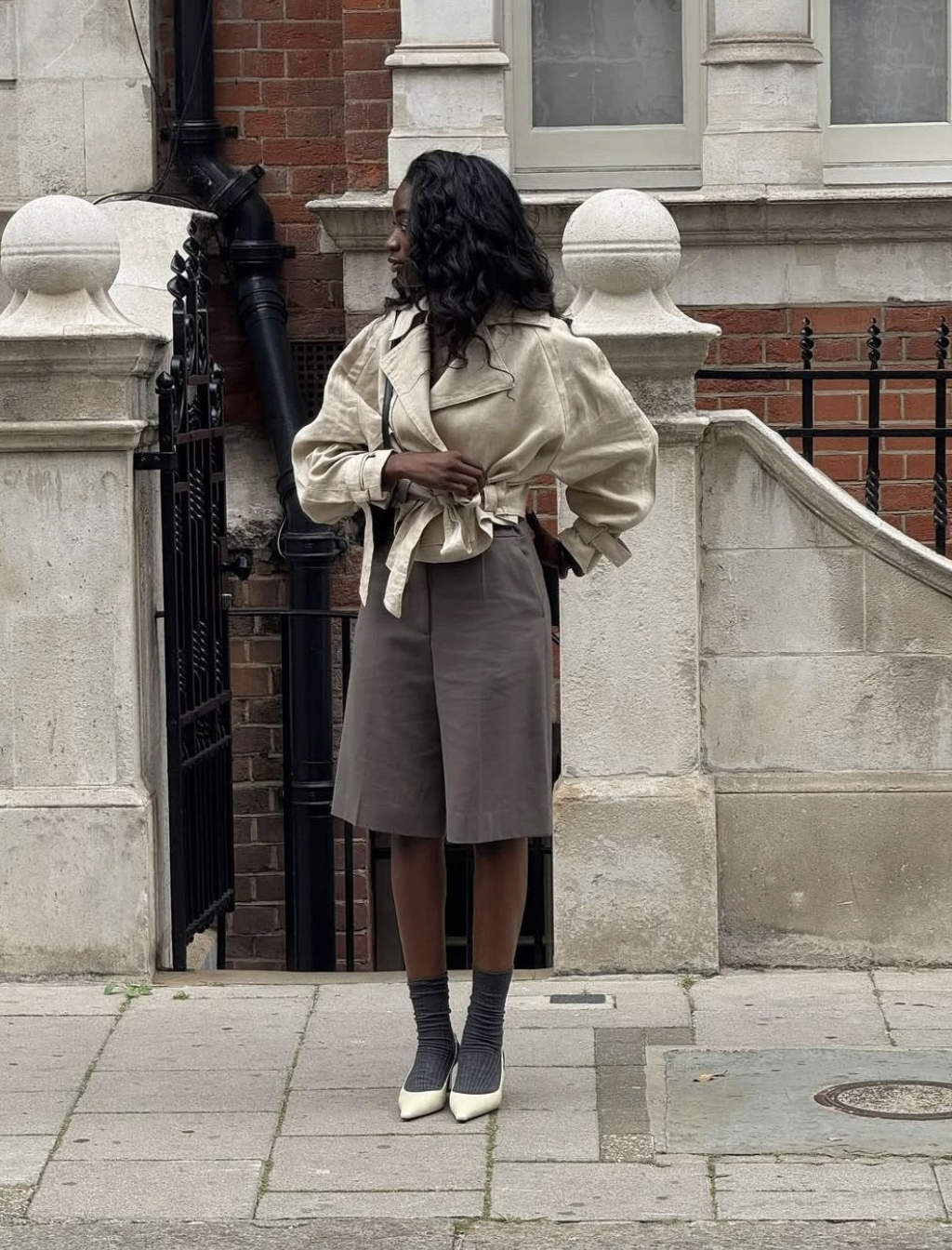 A woman wearing a neutral belted jacket and grey shorts paired with white slouchy socks and loafers, showcasing a classic poetcore aesthetic outfit.