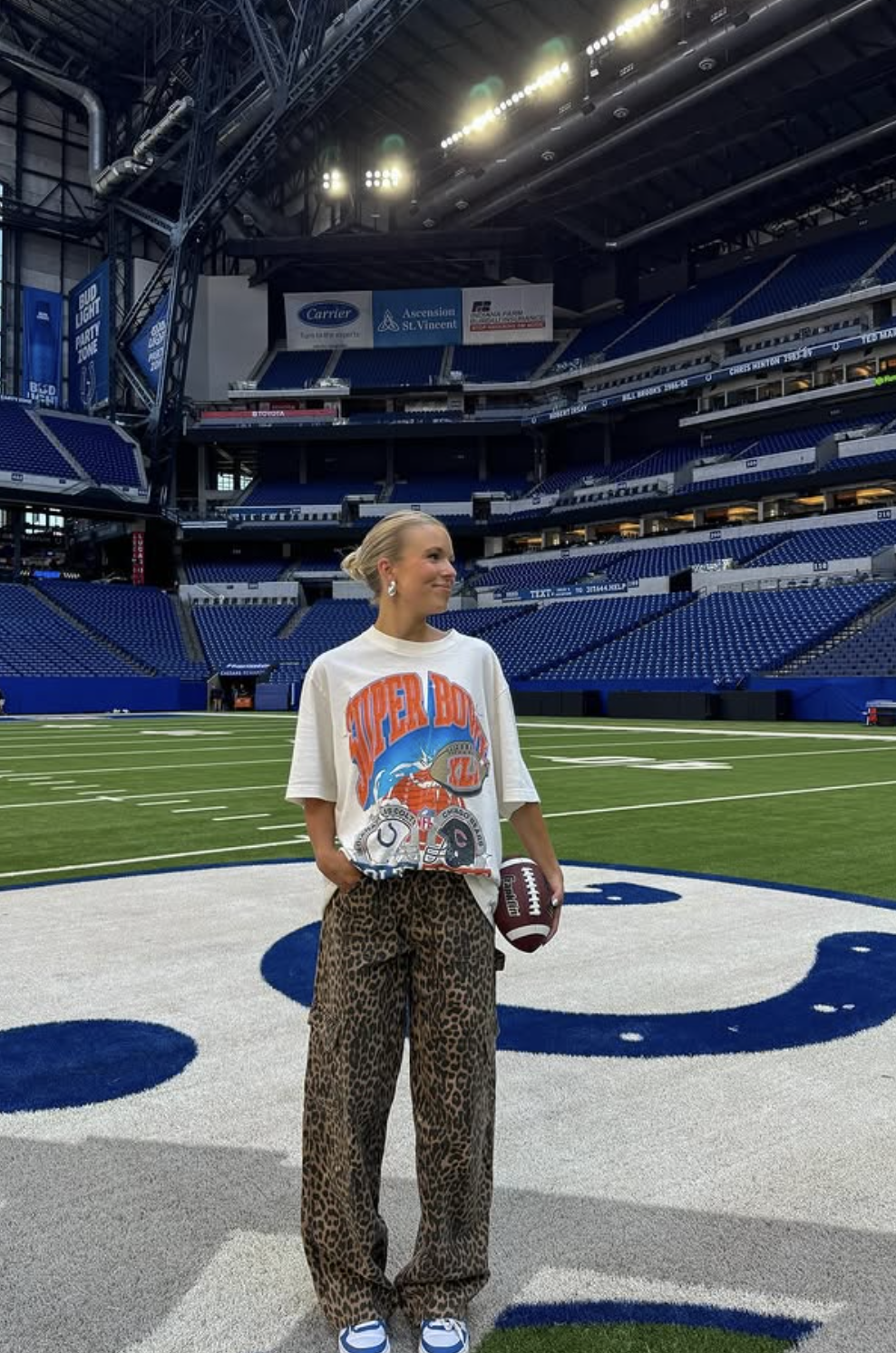 Woman on a football field wearing an oversized white football graphic tee, leopard-print wide-leg pants and blue-and-white sneakers.