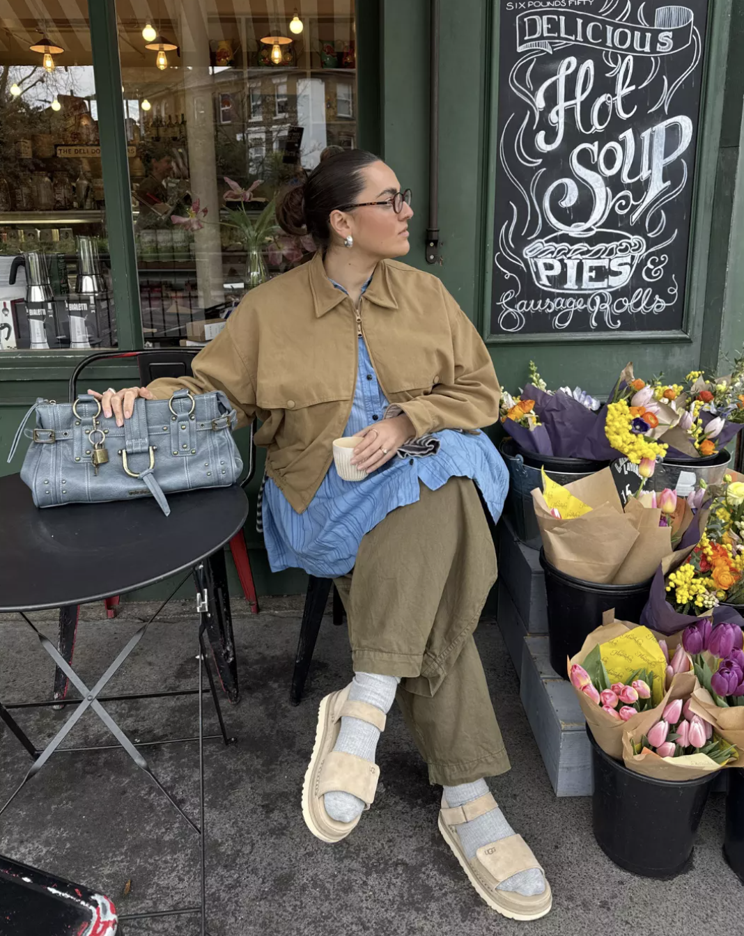 A woman sitting at an outdoor café table wearing a tan jacket, a blue button-down shirt, olive green relaxed trousers, light grey ribbed socks, and beige open-toe slide sandals, with a blue textured handbag resting on a black table.