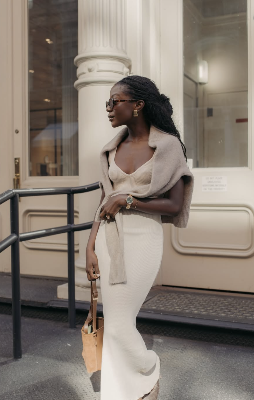 A woman wearing an elegant monochromatic cream maxi dress and a matching knit top, standing next to a white architectural column.